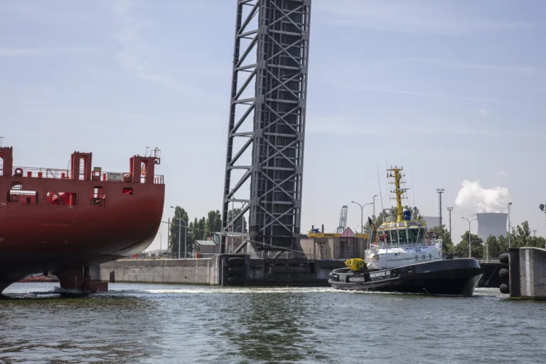 Harbour tug assisting a large vessel beside a lift bridge in an industrial port.